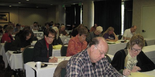 A room full of tables of men and women writing.