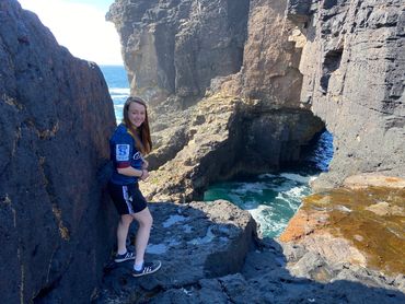 A young woman stands at a clifftop, overlooking a waterfall and a natural arch in the cliff beyond