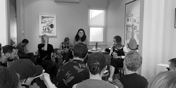  A  woman sits in a chair in front of a microphone in a room full of onlookers. 