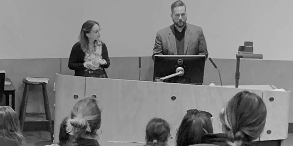 A woman and a man stand in front of a lecture hall delivering a speech. The woman holds a fidget toy