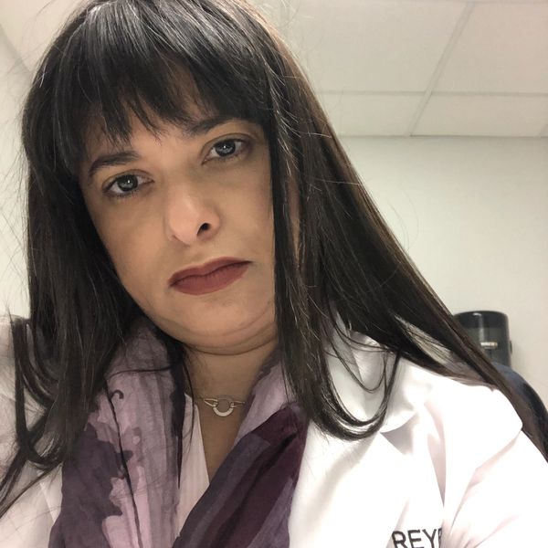 Woman with long dark hair and bangs wearing a white coat indoors.