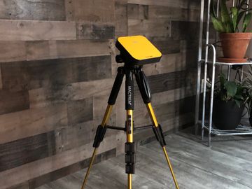 A yellow surveying tripod with a mounted device indoors on wooden flooring.