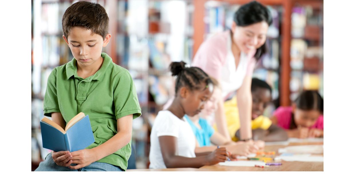 Elementary boy sits in library and looks sadly at a book. Classmates in background smile and reading