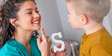Woman Speech Therapist sits with young boy. She holds letter "S" and points to her mouth to say "S" 