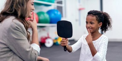 Young girl sits with woman. child holds mirror and finger to her lips during speech therapy