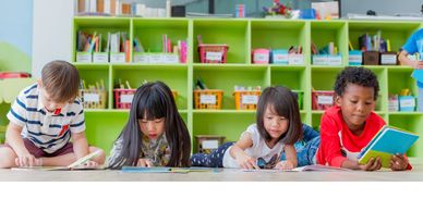 Four preschool age children lay on floor reading books. A bookcase behind them filled with books