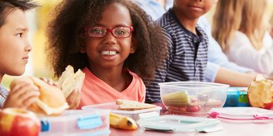 Group of children sit at cafeteria table smiling and eating sandwiches and fruit and vegetables