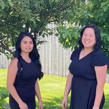 Two women with black hair smiling, wearing black dresses with trees in the background