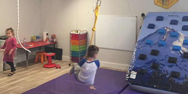Children playing on indoor gym equipment with mats and climbing wall.