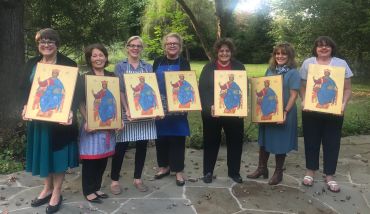 Seven women holding religious paintings outdoors, smiling.