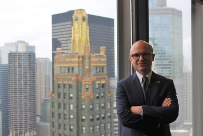 A man in a suit stands confidently by a window overlooking a cityscape with tall buildings.