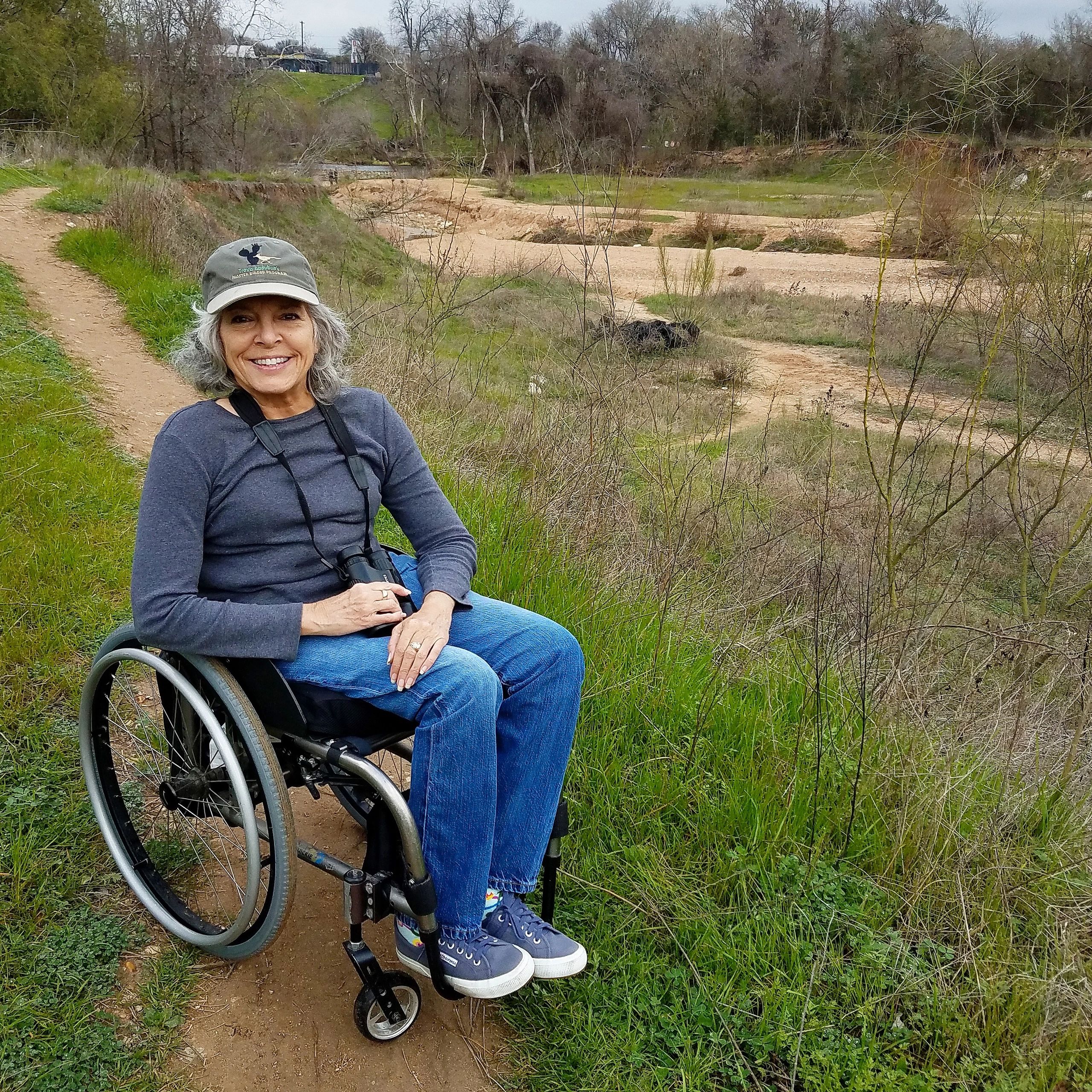 A woman with gray hair wearing a baseball hat, jeans, and gray shirt with binoculars around her neck