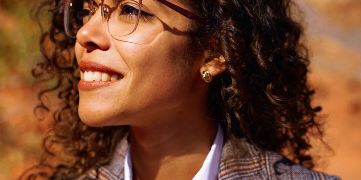Close-up of a smiling woman with curly hair and glasses in warm sunlight.