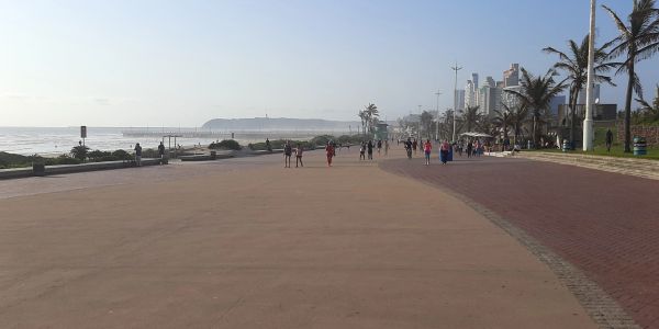 People walking along a beachfront promenade with city buildings in the background.
