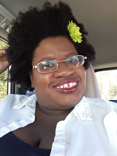 In Lyft, day, smiling mouth open showing gaps, afro with green flower, black tank, white button down