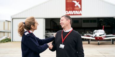 Dos personas se saludan frente a un hangar de aviones.