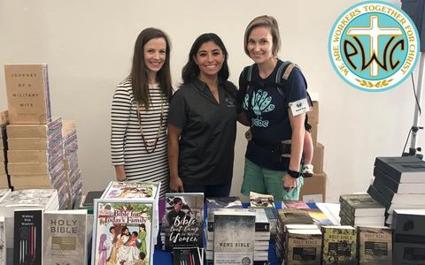 Three women stand behind a table filled with books at an event.