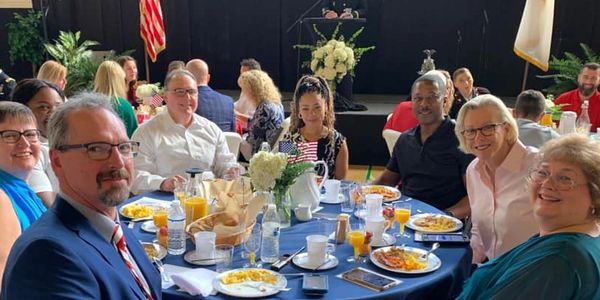 Group sits around table with food and drinks, smiling at camera.