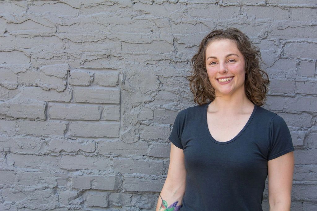 Colour photo of Meagan smiling in front of a grey brick wall, grey tshirt, hummingbird tattoo
