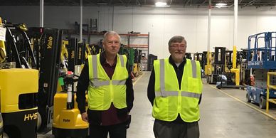 Two men in yellow safety vests standing in a warehouse with forklifts.