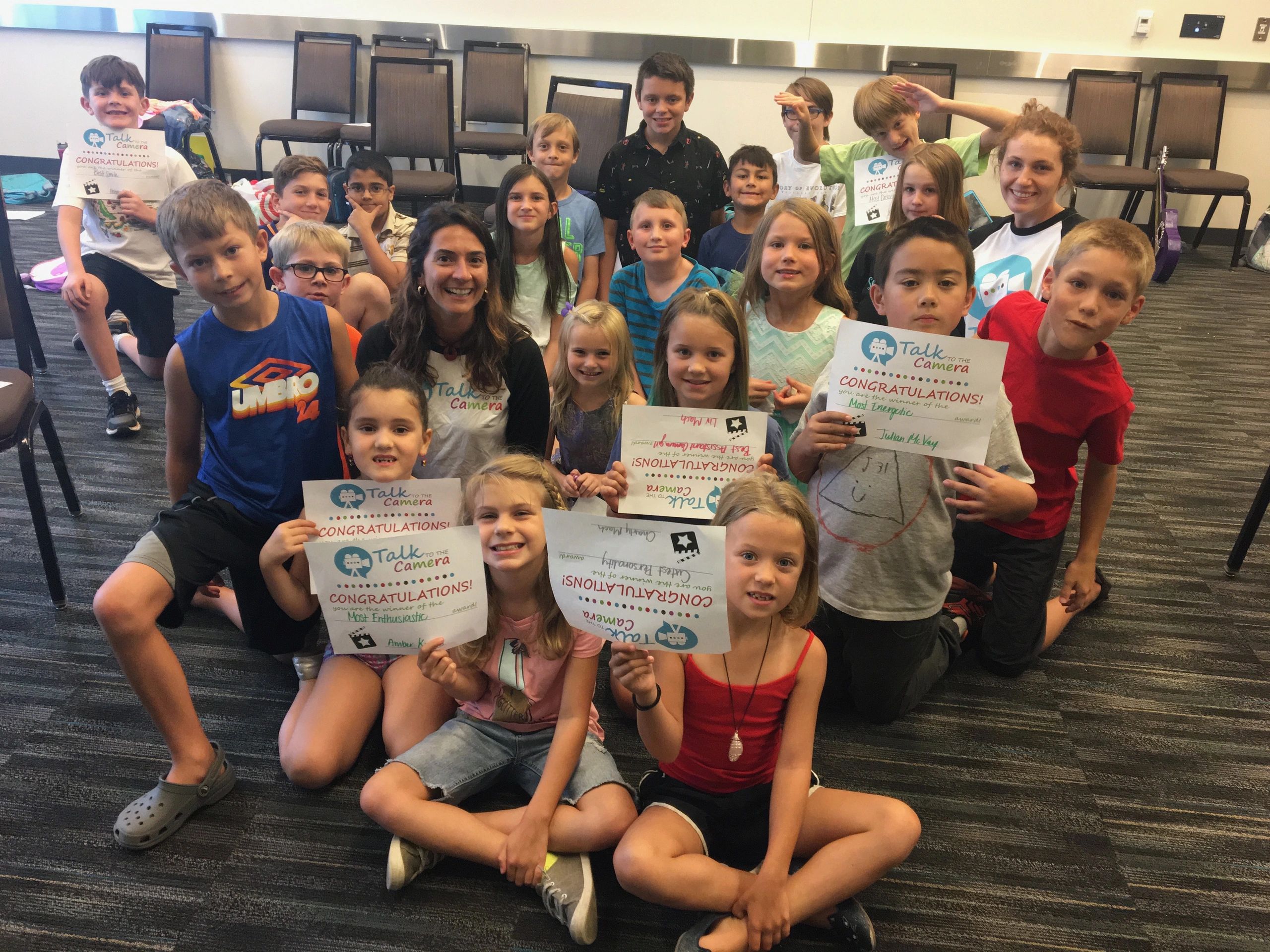 Students and teachers sitting in a classroom floor, smiling and holding up their class certificates