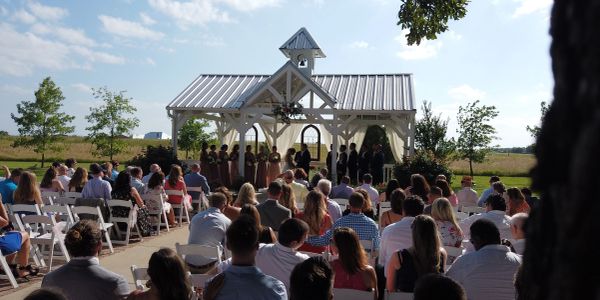 Outdoor wedding ceremony with guests and bridal party under a pavilion.