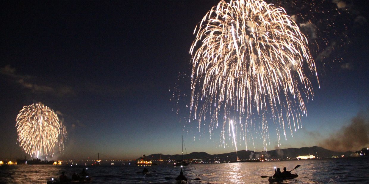 Fireworks light up the night sky over a body of water with silhouetted kayakers.