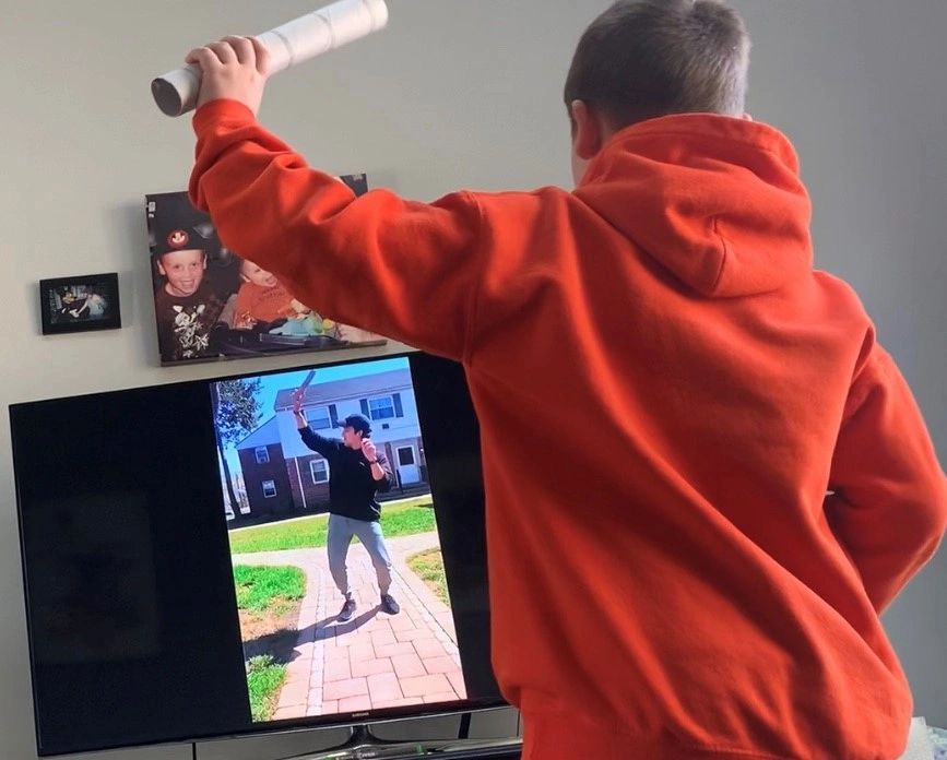 A boy in an orange hoodie mimics a person on TV holding a similar object.