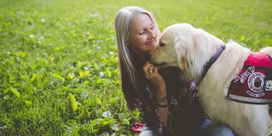 Joey Ramp sitting on a patch of green grass smiling while her service dog Sampson is looking towards