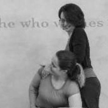 Two women interacting in a grayscale photo with one woman standing behind the other.