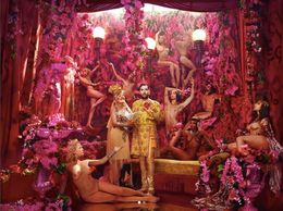 A man in ornate attire surrounded by pink-clad dancers in a lavish floral setting.