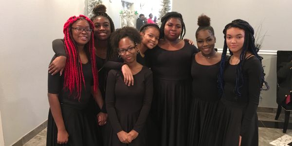 Seven young women in black dresses posing together indoors.