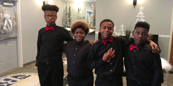 Four boys dressed in black shirts and red bow ties posing indoors.