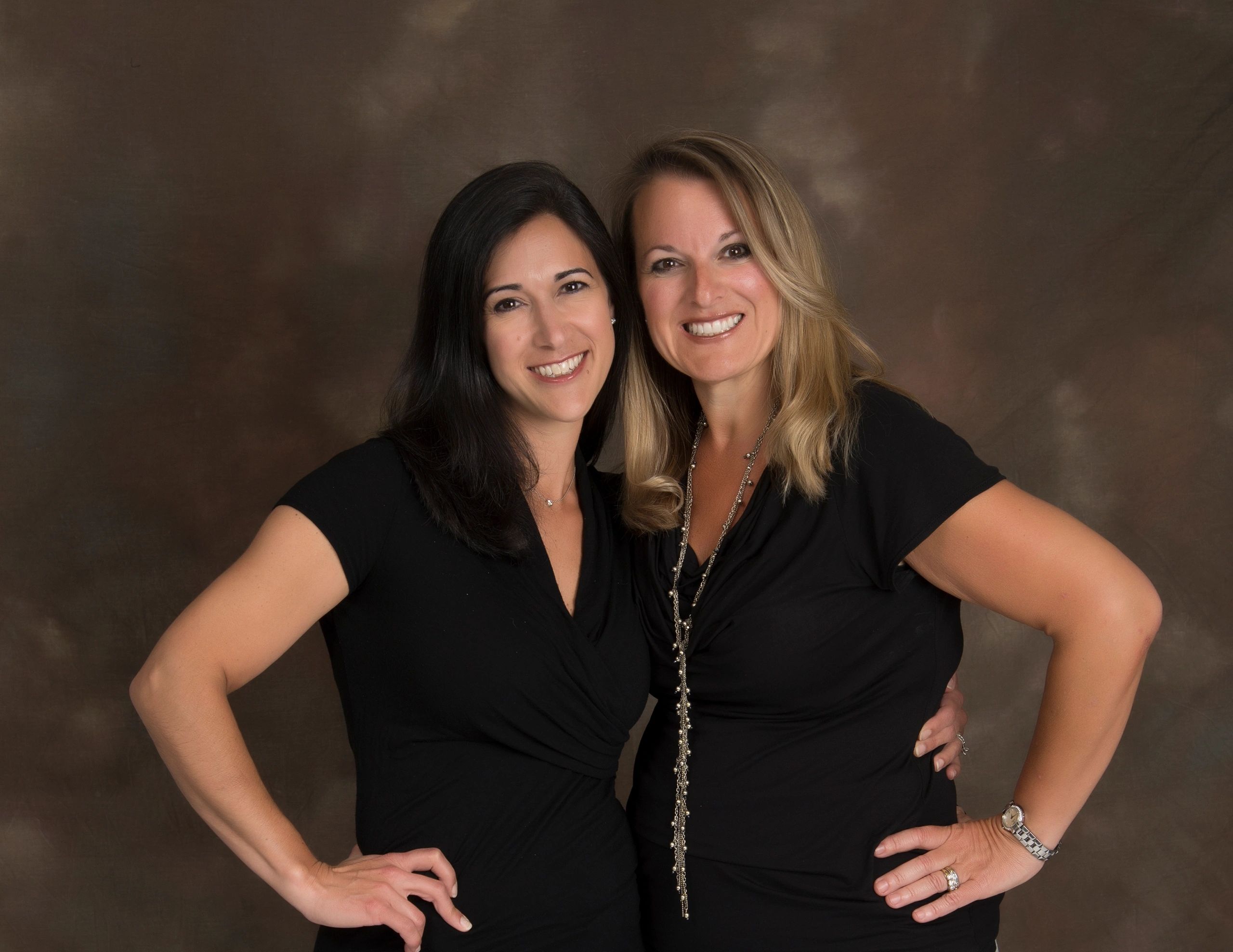 Two smiling women posing together with arms around each other against a brown backdrop.