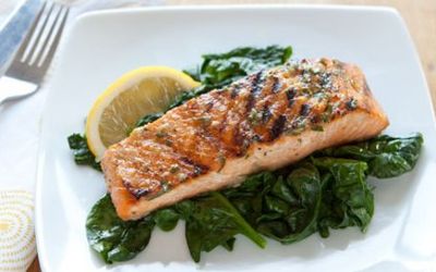 A white plate with a bed of sauteed spinach and a piece of grilled salmon and fresh lemon on top