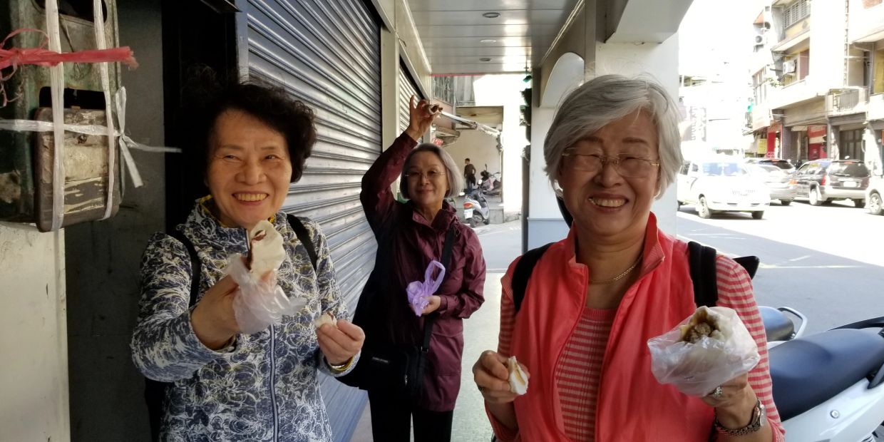 Three elderly women smiling and holding food on a sunny street.