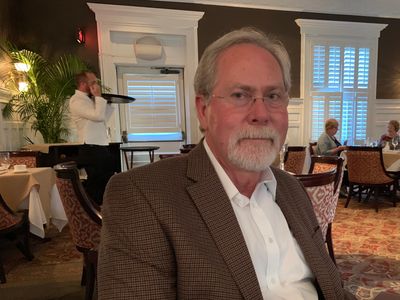 An elderly man with glasses and a beard sits in a restaurant holding a menu.