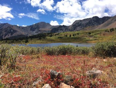 High rockies mountain range, blue sky, white fluffy clouds, lake and red foliege brush in foreground.