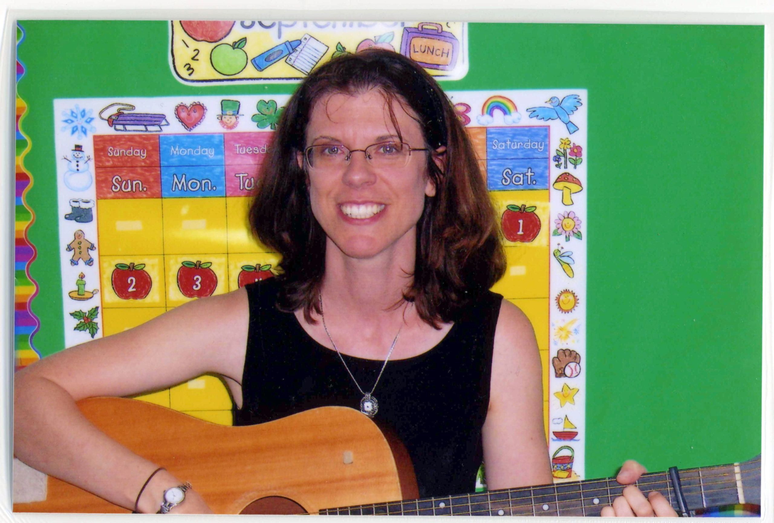 Picture of Margie with a guitar in front of a bulletin board. 