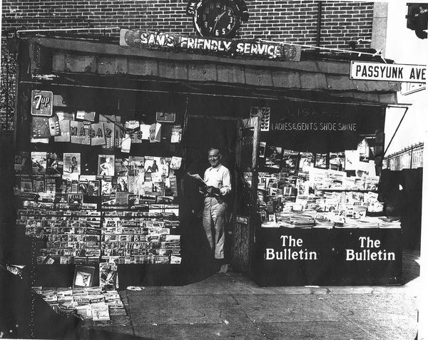 Black and white picture of man standing in front of his newsstand on street corner