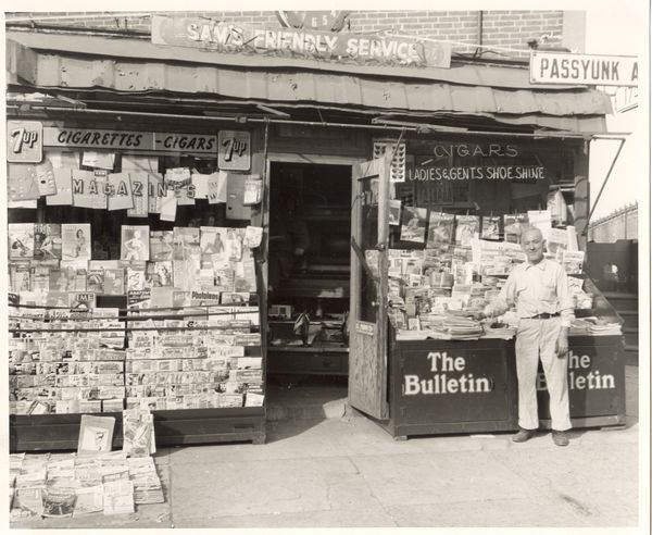 Black and white picture of man in front of newsstand with magazines on street corner