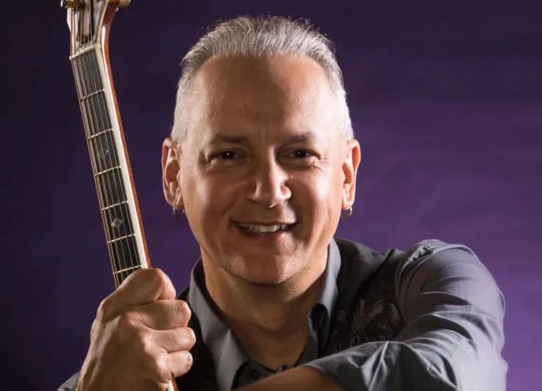 Smiling man holding a guitar neck against a purple background.
