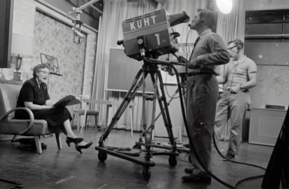 Black-and-white photo of a TV studio with a woman seated and two men operating cameras.