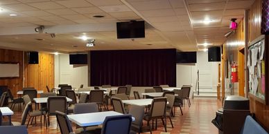 Empty community hall with rows of tables and chairs facing a stage.