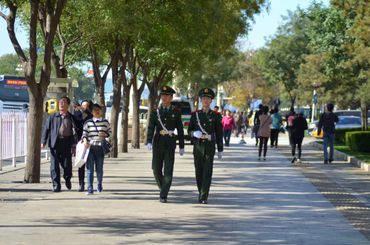 Two uniformed officers walk down a tree-lined urban sidewalk with pedestrians around.