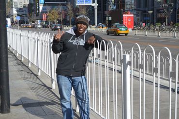 A person wearing a black hoodie and beanie flashes a peace sign on a city sidewalk.