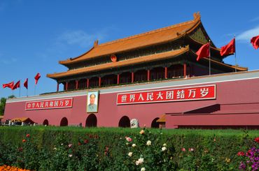 The Tiananmen Gate with red flags and Mao Zedong portrait under a clear blue sky.