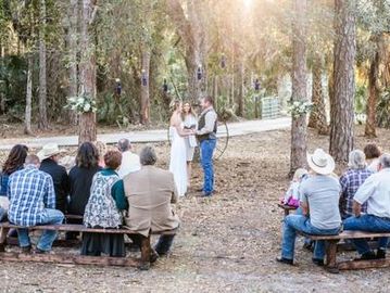 Outdoor rustic wedding ceremony in a wooded area with guests seated on benches.