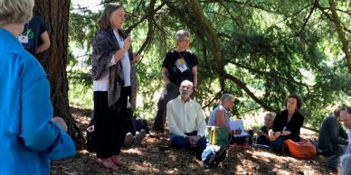Group gathered outdoors under trees, one person speaking.