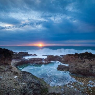 Sunset over a rocky coastline with crashing waves.